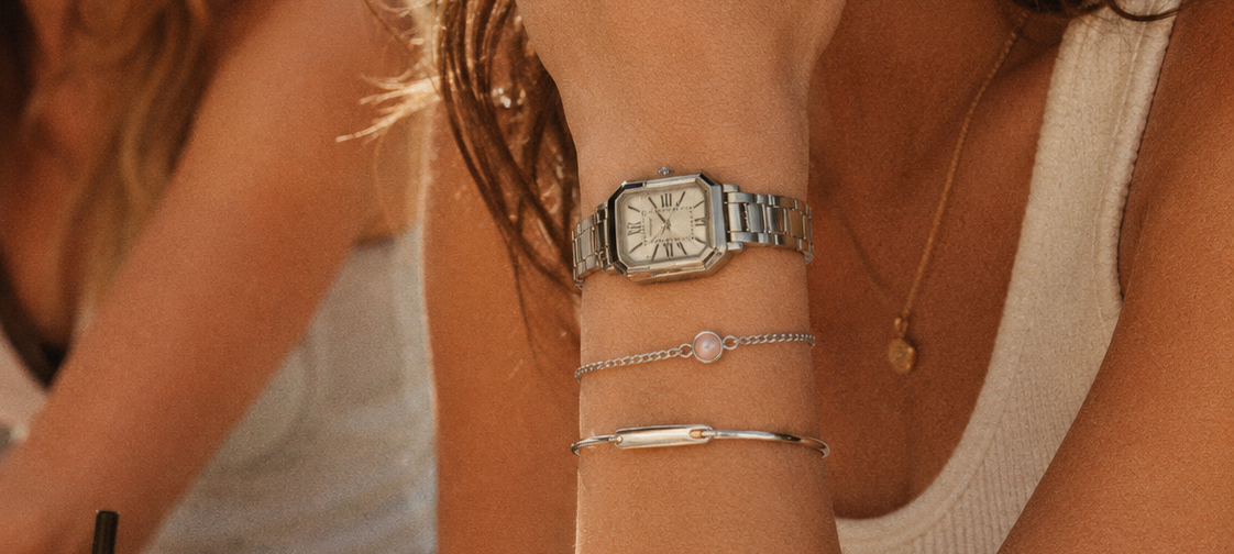 Woman wearing a watch and bracelets with a blurred background of another person.