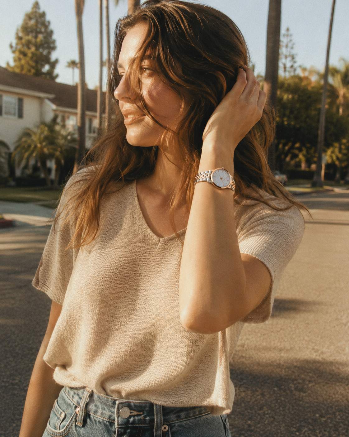 A woman wearing a beige short-sleeve knit top and blue jeans, standing outdoors on a sunny street with palm trees and houses in the background. She is wearing a silver-toned watch on her wrist.