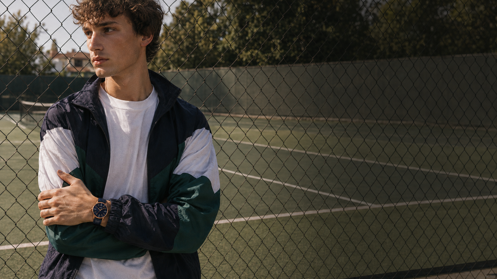 A man standing on a tennis court behind a chain-link fence, wearing a navy, white, and green colorblock windbreaker jacket and a brown leather watch.