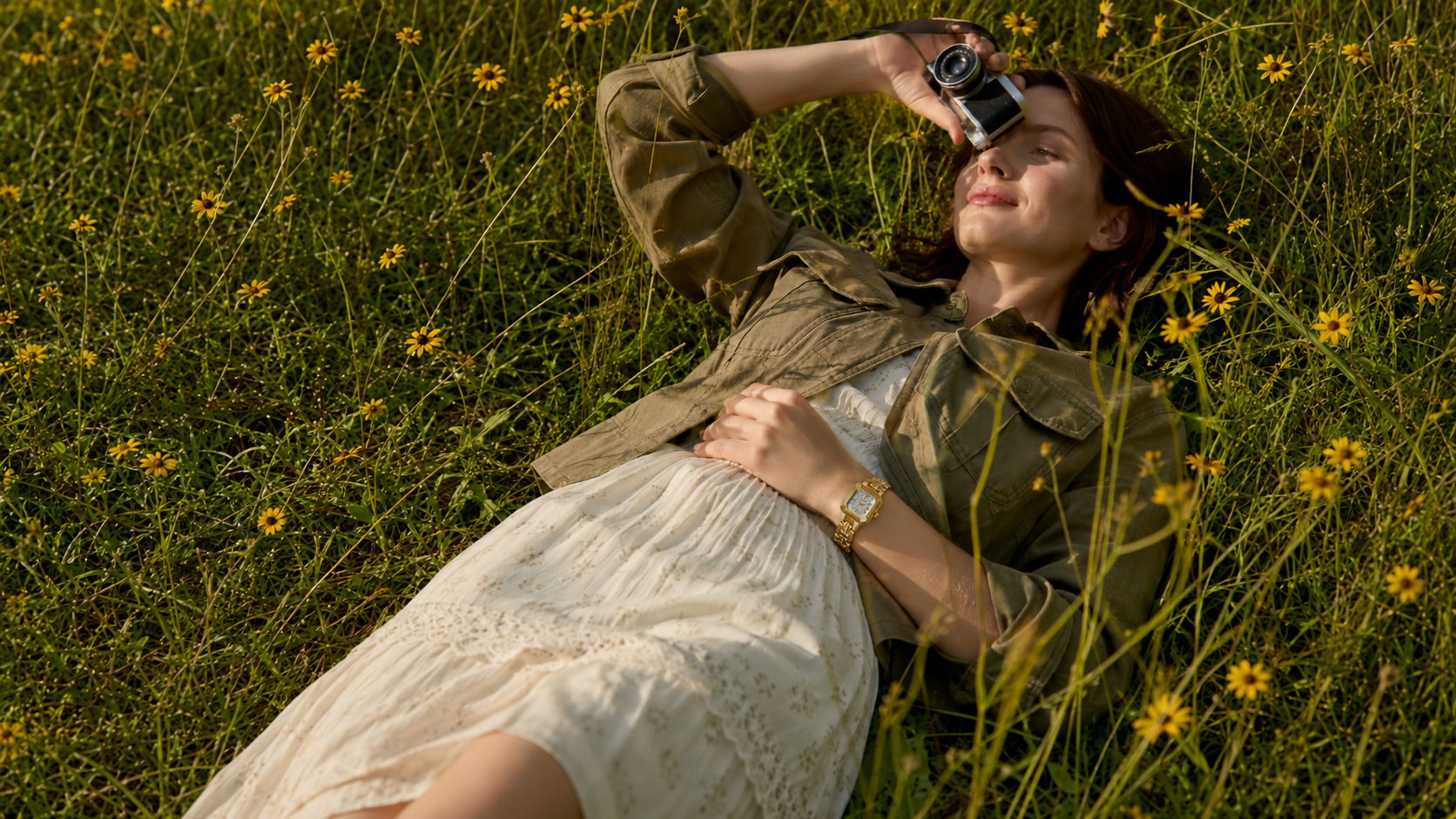 A woman lying in a grassy field of small yellow flowers, wearing an olive green jacket and a white dress, holding a camera to her eye.